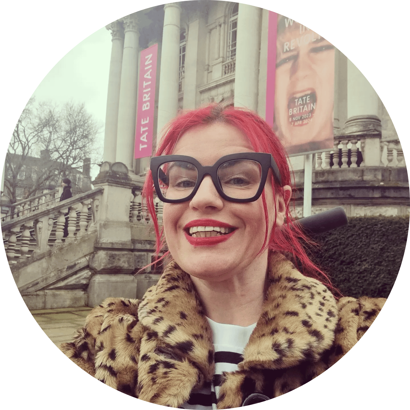 A picture of Kate Annetts, a white woman with thick black glasses and dyed pillar box red hair.